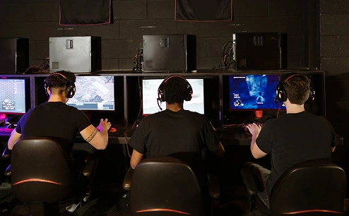E-sport játékosok hátulnézete, akik PC-n játszanak csapatkörnyezetben, több monitorral és headsetekkel.